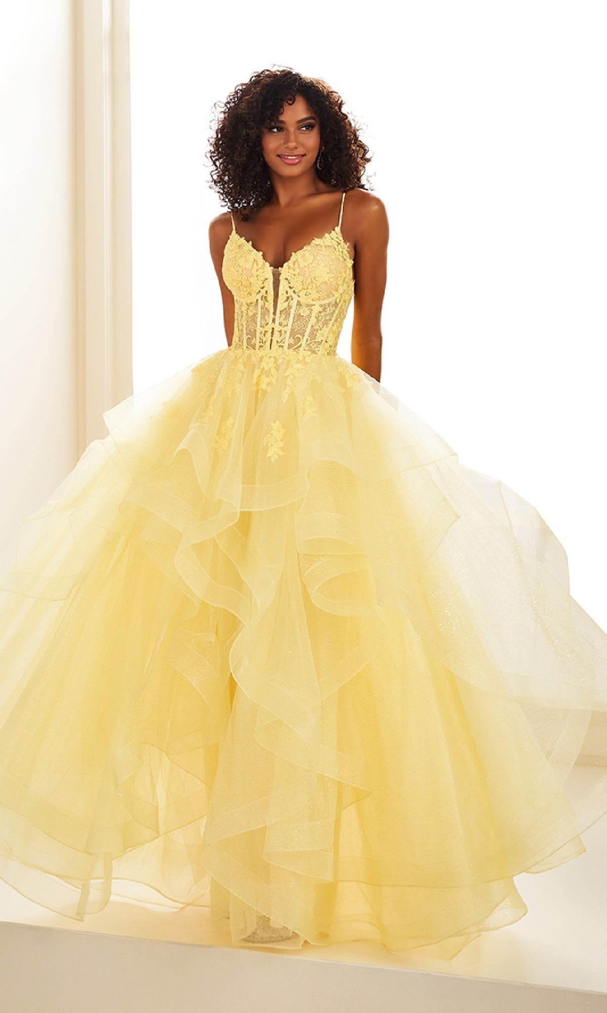 A woman with curly hair models the Ellie Wilde EW36203 Glitter Tulle Prom Ball Gown, featuring a yellow ruffled skirt, embroidered corset bodice, and thin straps, standing against a white background.